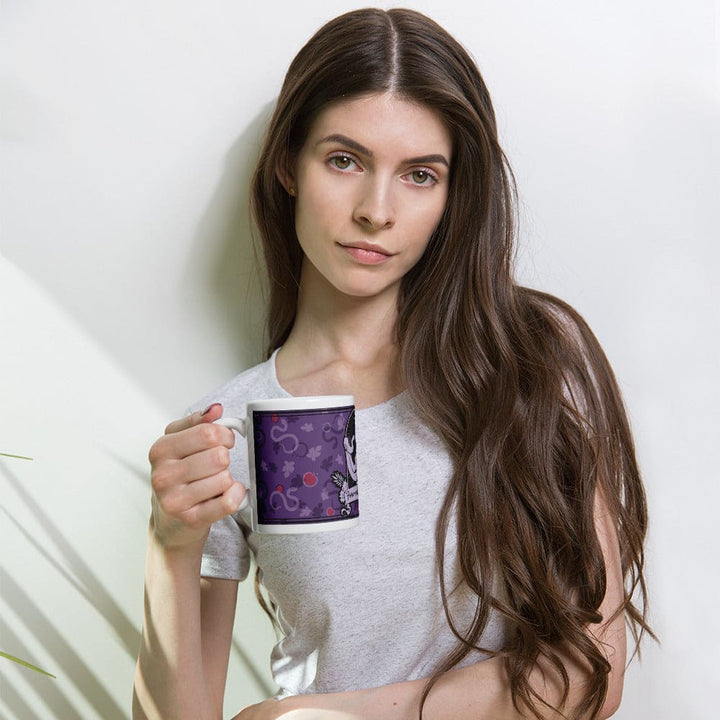 A white ceramic mug featuring a purple background with a repeating pattern of small red apples and gray snake-like shapes. A woman holds the mug while leaning against a white wall.