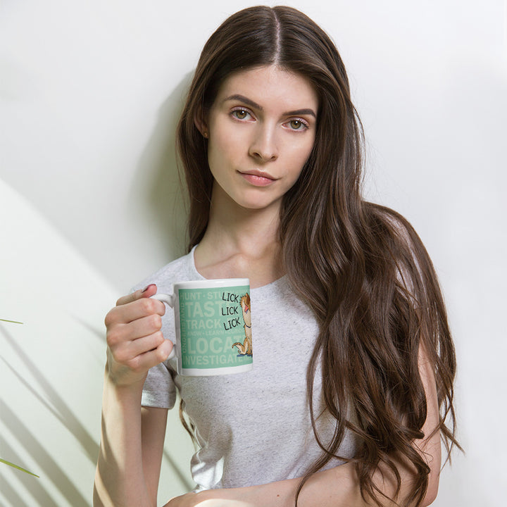 A white ceramic mug featuring a light green background with the text HUNT, TASTE, TRACK, LOCATE, INVESTIGATE and the repeated word LICK next to an illustration of a bearded dragon lizard. The mug has a curved handle and a smooth, glossy finish.