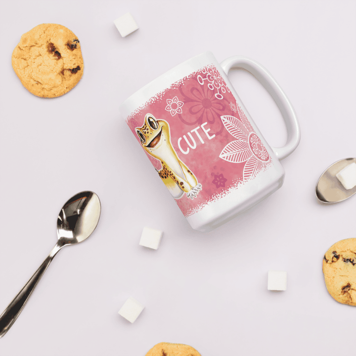 A white ceramic mug with a pink floral design and a cartoon leopard gecko, surrounded by cookies, sugar cubes, and a spoon on a light pink background.