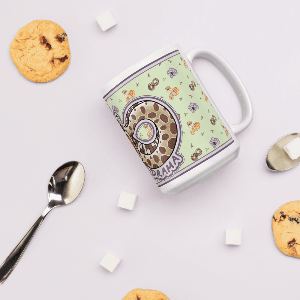 A white ceramic mug featuring a light green background with a repeating pattern of small colorful snakes and a large central graphic of a coiled snake with the text "DRAMA" printed in purple letters on the side of the mug. It sits on a table with sugar cubes and cookies  and a spoon surrounding it.