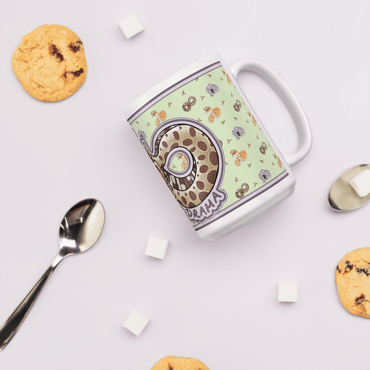 A white ceramic mug featuring a light green background with a repeating pattern of small colorful snakes and a large central graphic of a coiled snake with the text "DRAMA" printed in purple letters on the side of the mug. It sits on a table with sugar cubes and cookies  and a spoon surrounding it.