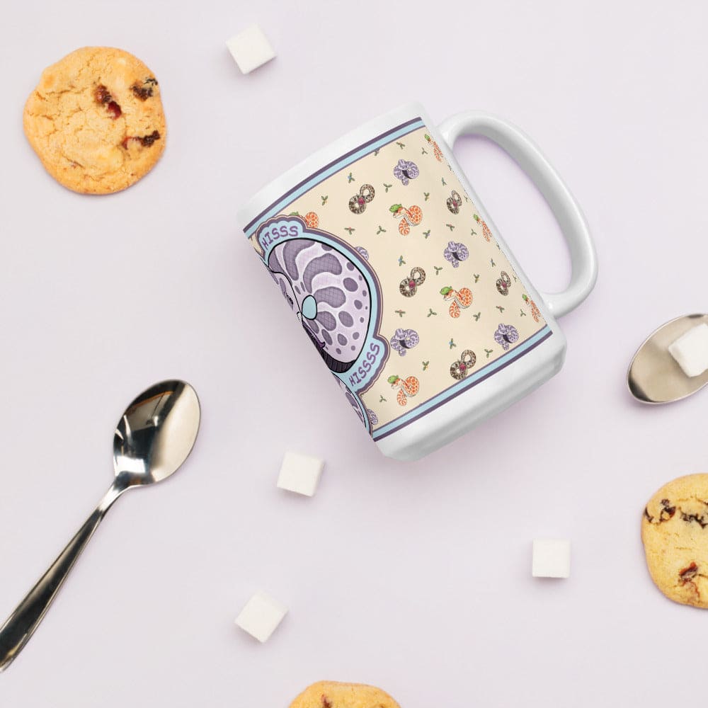 Mug with a design of a purple hognose snake with some text saying "HISS" around the animal. The mug is resting on it's side showing the left side of the design, which is a soft yellow with tiny hognose snakes and flowers in a vintage print and surrounded by cookies and spoons on a light background