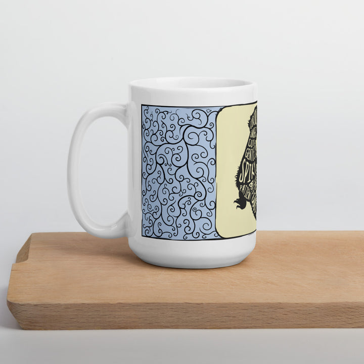 A white ceramic mug featuring a graphic design with a light blue section covered in black swirling patterns and a pale yellow section containing a black silhouette of a bearded dragon filled with various words including the word spiky. The mug sits on a wood countertop with a white background.