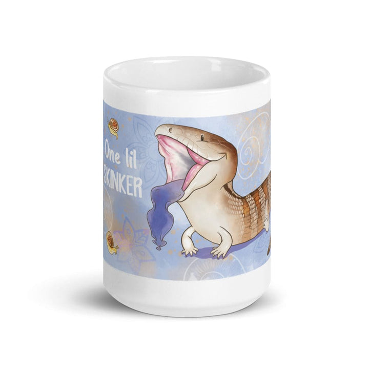 A white 15oz ceramic mug featuring a cartoon illustration of a blue-tongued skink with its mouth open, displaying a bright blue tongue. The background is light blue with small snail graphics and the text One lil SKINKER printed in white. The mug sits on a blank white background.