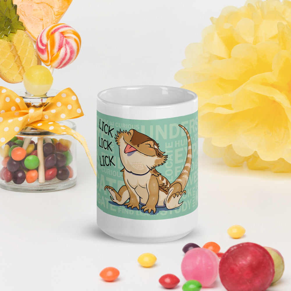 A white ceramic mug with a cartoon lizard illustration and the text 'LICK LICK LICK' on a mint green background, styled on a white surface with candy, a yellow flower, and a lollipop jar in the background.