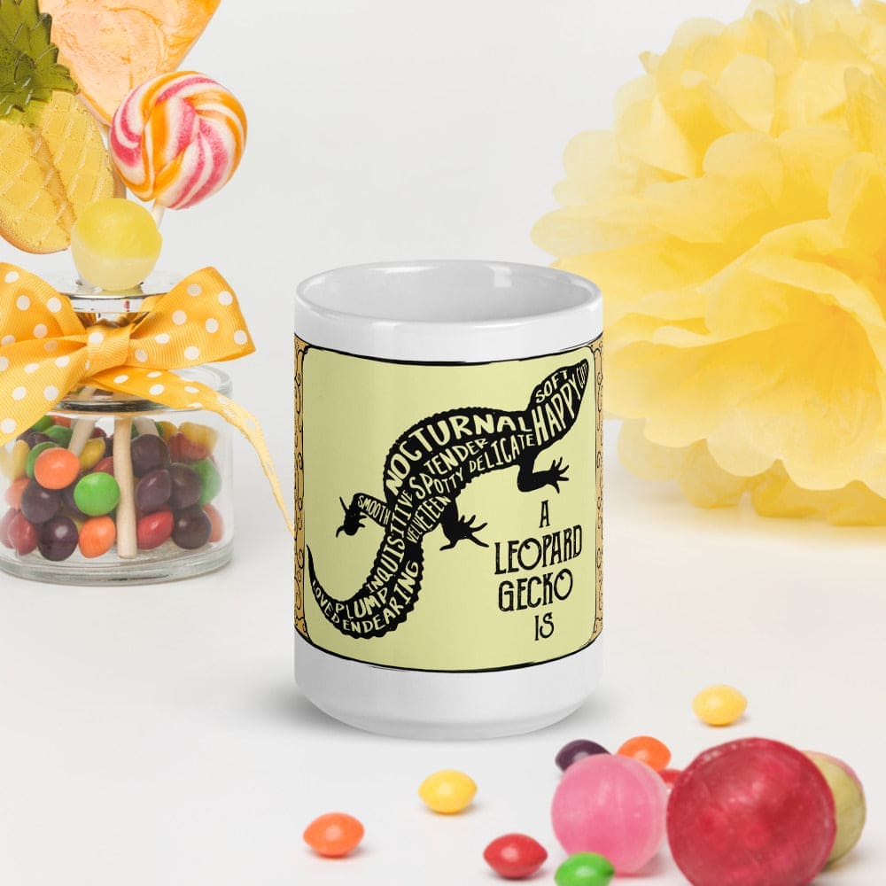 A yellow ceramic mug with a silhouette of a leopard gecko with text inside it's body, resting on a white surface surrounded by candy and bright colored flowers.