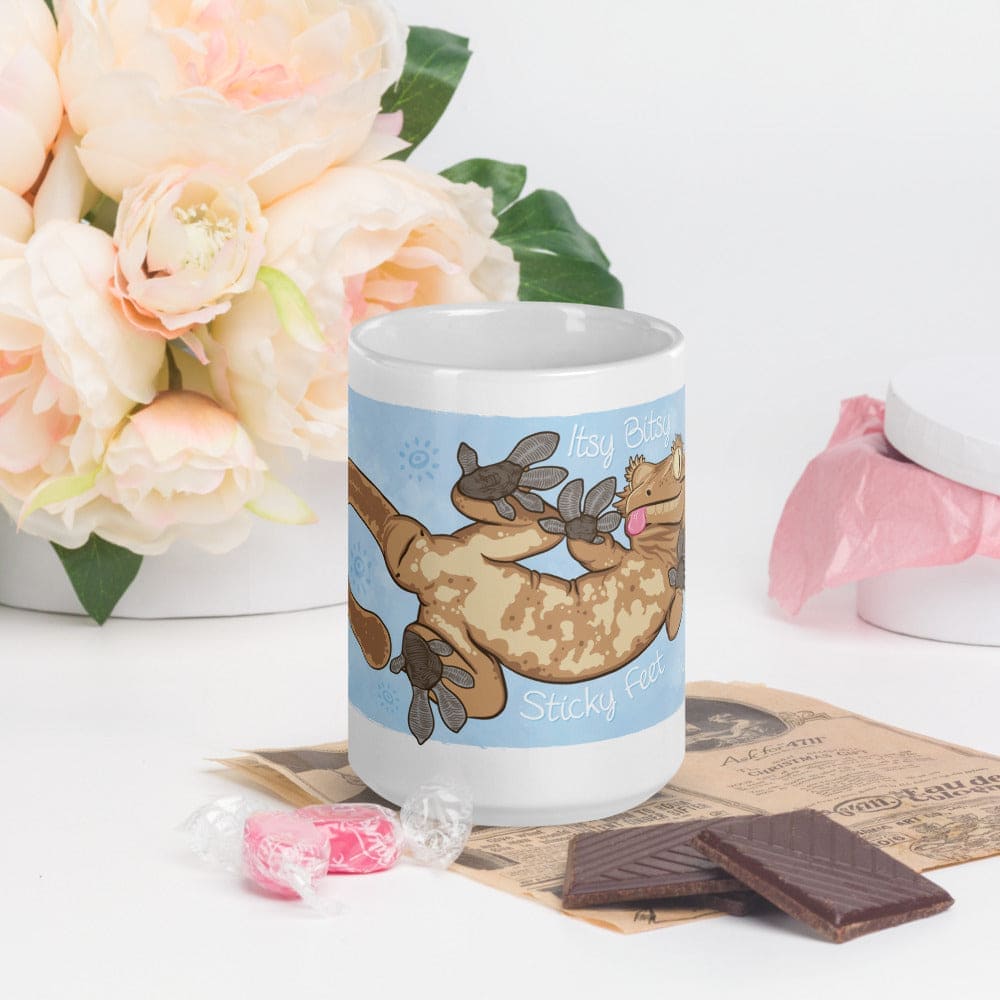 A white ceramic mug featuring a cartoon illustration of a tan and brown crested gecko with its belly and feet pressed against a light blue background. The text Itsy Bitsy Sticky Feet is printed in white script around the gecko.
