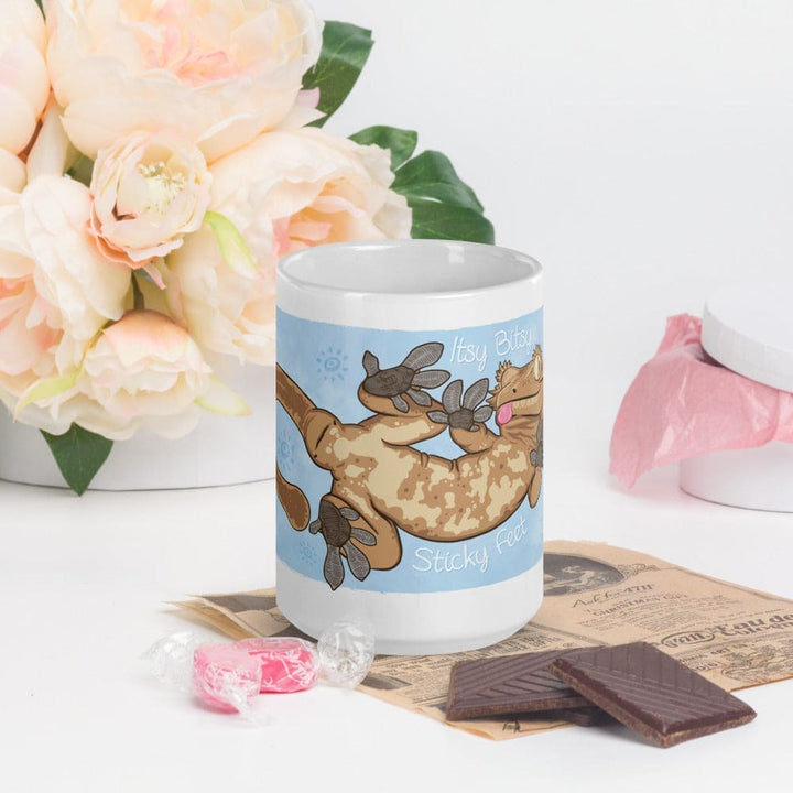 A white ceramic mug featuring a cartoon illustration of a tan and brown crested gecko with its belly and feet pressed against a light blue background. The text Itsy Bitsy Sticky Feet is printed in white script around the gecko.