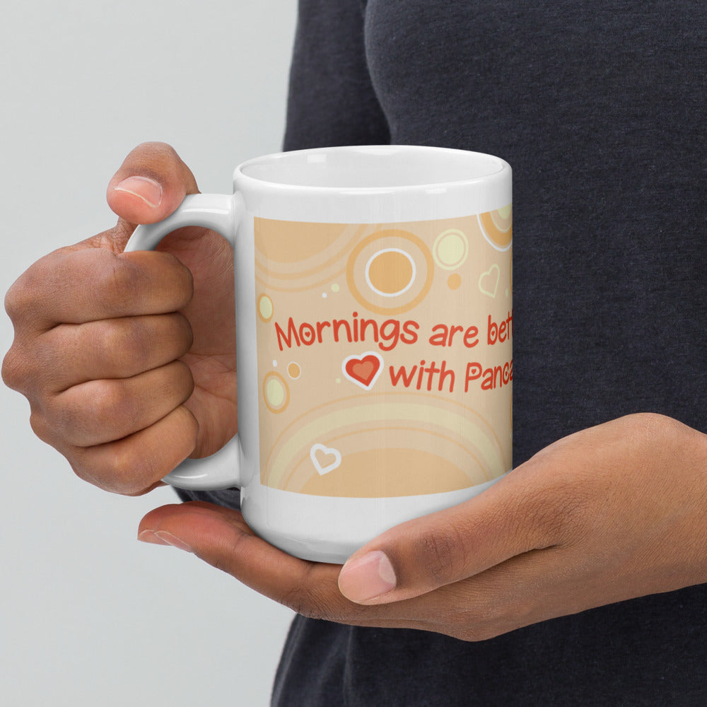 Hands holding a white ceramic mug featuring a light orange background with concentric circles and small heart shapes. The text printed on the mug reads, "Mornings are better with Pancakes," with a small red heart icon between the words.