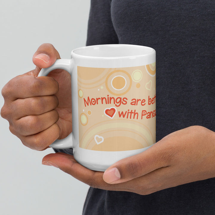 Hands holding a white ceramic mug featuring a light orange background with concentric circles and small heart shapes. The text printed on the mug reads, "Mornings are better with Pancakes," with a small red heart icon between the words.