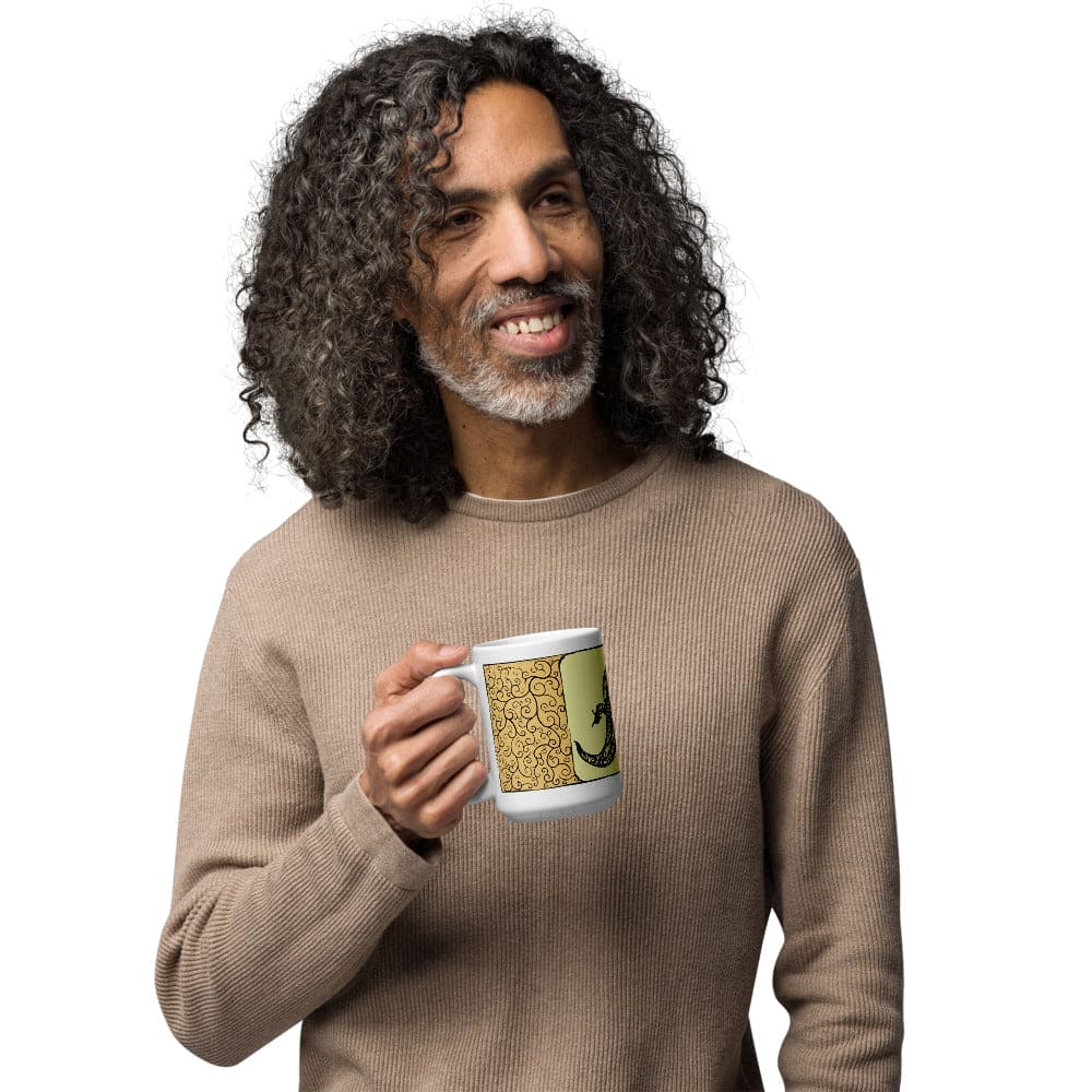  A man holding a white ceramic mug featuring a decorative panel with a yellow background, intricate black swirling filigree patterns, and a black silhouette of a lizard. The mug has a standard handle and a cylindrical shape for holding beverages.