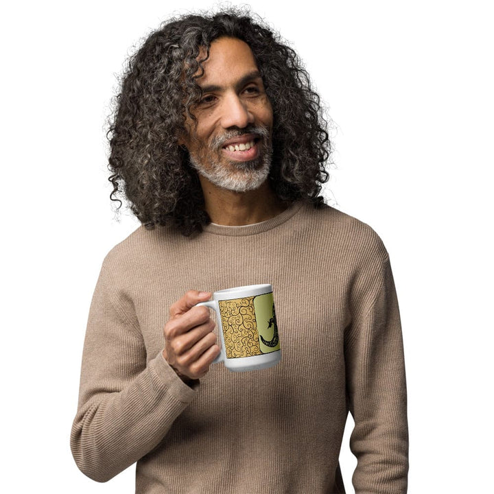  A man holding a white ceramic mug featuring a decorative panel with a yellow background, intricate black swirling filigree patterns, and a black silhouette of a lizard. The mug has a standard handle and a cylindrical shape for holding beverages.