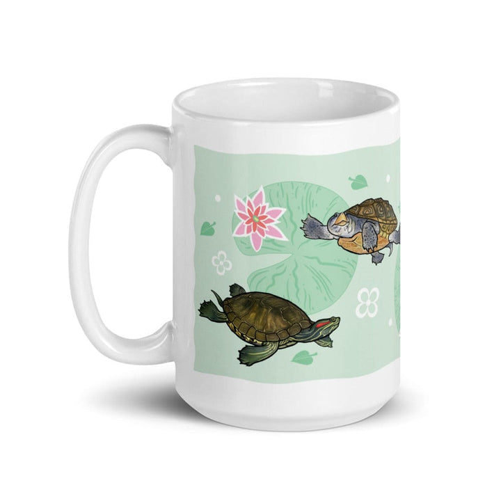 A white ceramic mug featuring a printed design of two turtles on a light green background with a pink lotus flower and lily pad motifs. The handle is on the left side of the mug.