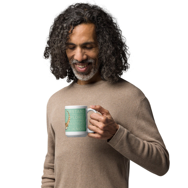 A man with long curly hair and a beard, wearing a beige ribbed sweater, holding a white ceramic mug with a mint colored wrap with multiple words printed on it. The background is solid white.