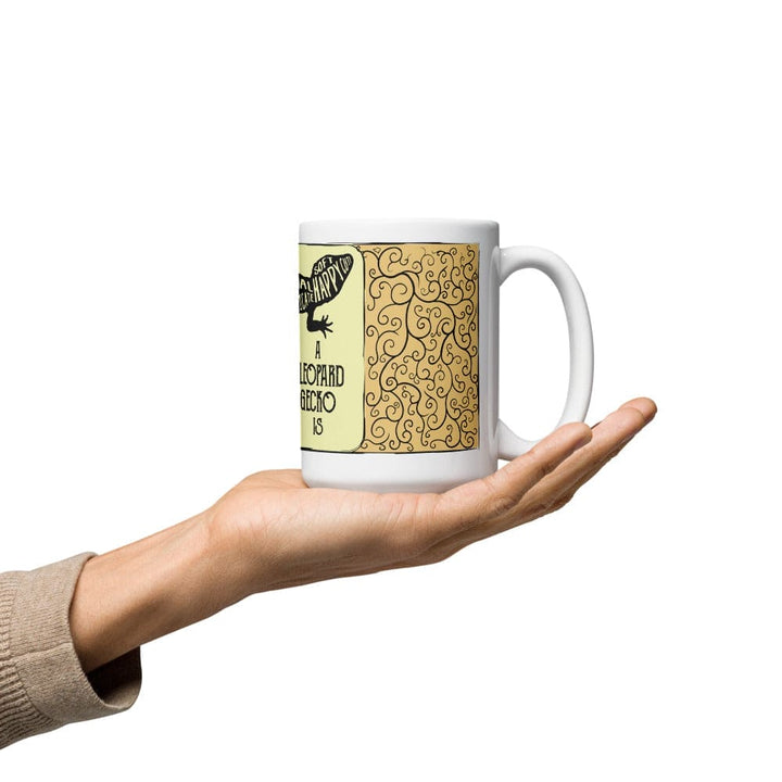 A  white ceramic mug in the palm of an outstretched hand featuring a black graphic of a leopard gecko and the text "A LEOPARD GECKO IS. The design includes a soft yellow panel with a black swirling vine pattern next to the gecko illustration and text.