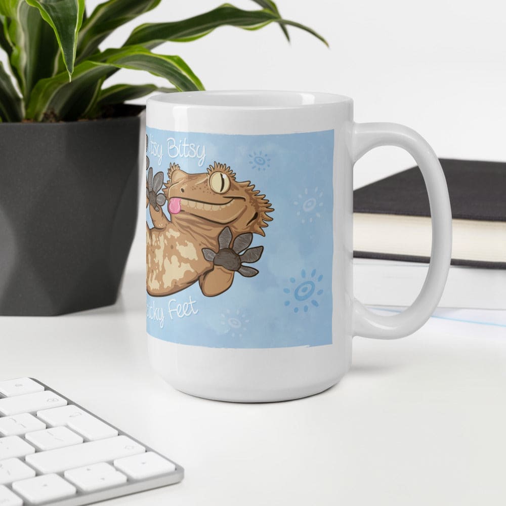 A white ceramic mug featuring a cartoon illustration of a tan crested gecko with its tongue out. The gecko is pressed against a light blue background with small circular patterns. The text reads Itty Bitty Sticky Feet. The mug sits on a white table near a keyboard and a potted plant.
