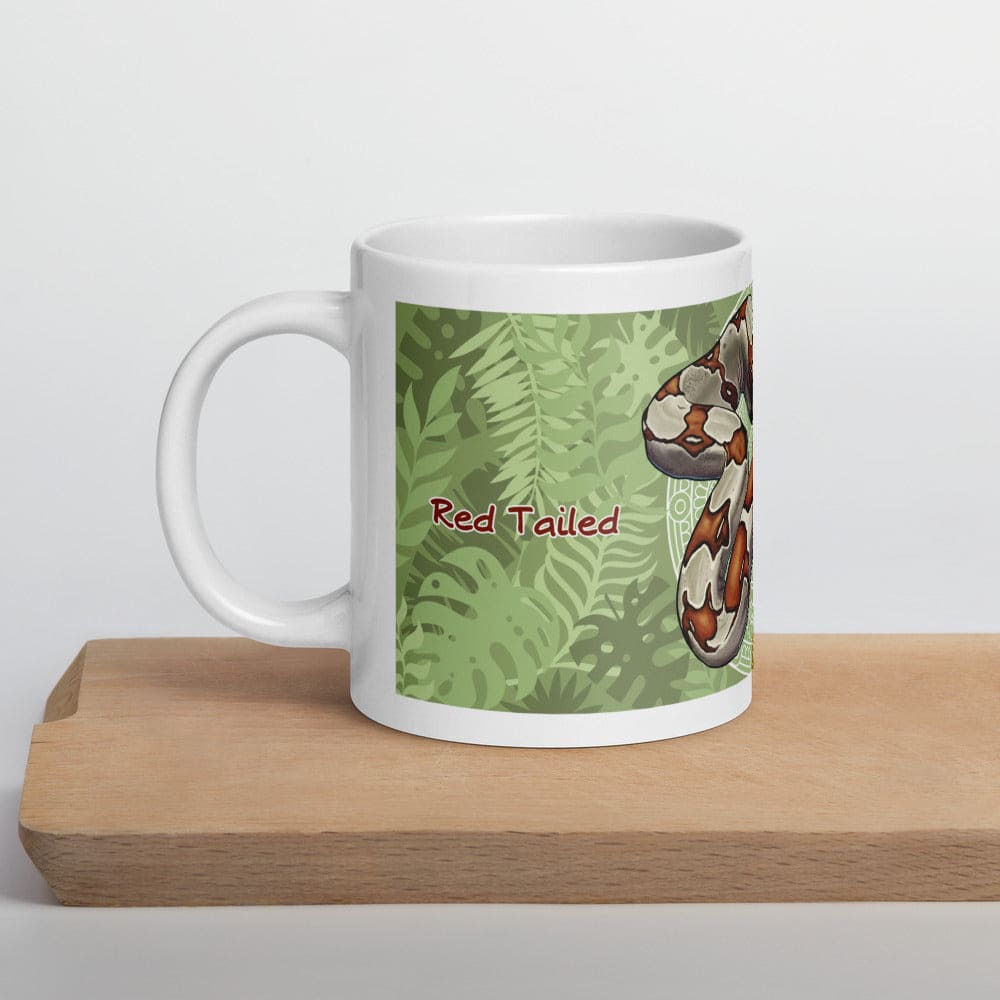 A white ceramic mug featuring a graphic illustration of a red-tailed boa constrictor against a green tropical leaf background. The text "Red Tailed" is printed in a dark red font on the side of the mug, which has a handle.