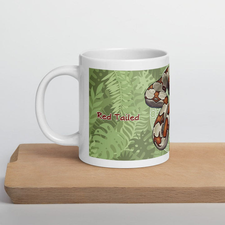 A white ceramic mug featuring a graphic illustration of a red-tailed boa constrictor against a green tropical leaf background. The text "Red Tailed" is printed in a dark red font on the side of the mug, which has a handle.