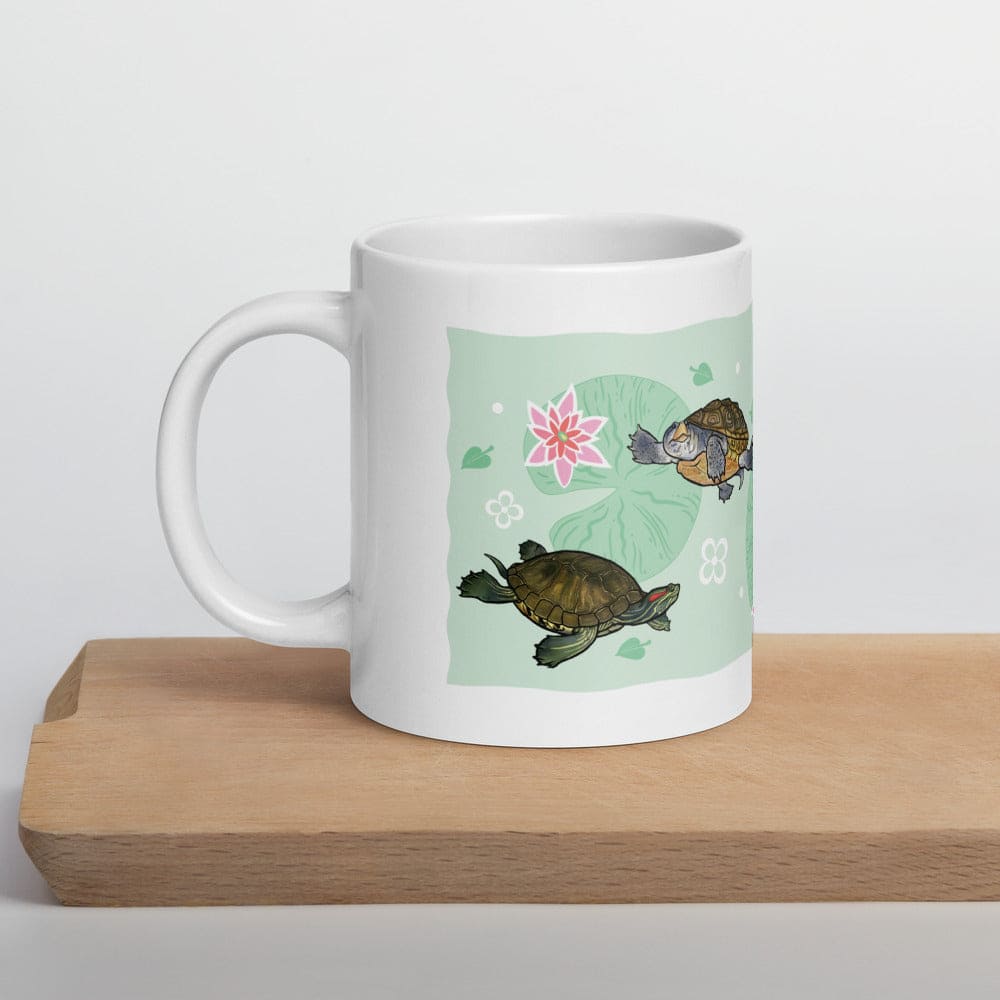 A white ceramic mug with a printed illustration of two turtles on a light green background, resting on a wooden cutting board against a white studio backdrop.