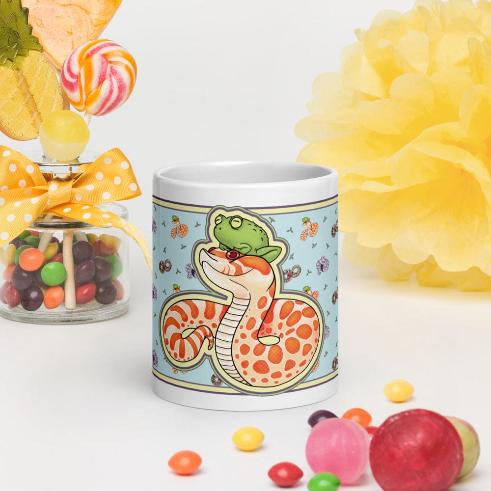 Image of a 11oz white ceramic mug with a graphic showing an albino hognose snake with a frog on it's head. The background color is blue with lots of other hognose snakes and flowers surrounding it. This is showing the front side of mug and is surrounded by colorful candies to eccentricate the cute design of the mug.