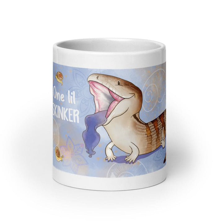 A white 20oz ceramic mug featuring a cartoon illustration of a blue-tongued skink with its mouth open, displaying a bright blue tongue. The background is light blue with small snail graphics and the text One lil SKINKER printed in white. The mug sits on a blank white background.