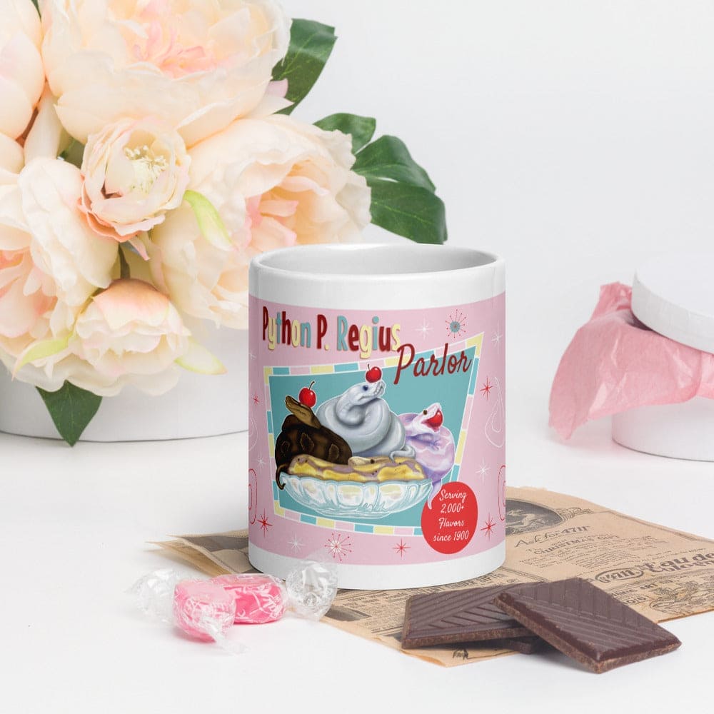 A ceramic mug depicting a retro style ball python design of snakes in a ice cream sundae bowl resting on a table surrounded by chocolate, flowers, and candy.