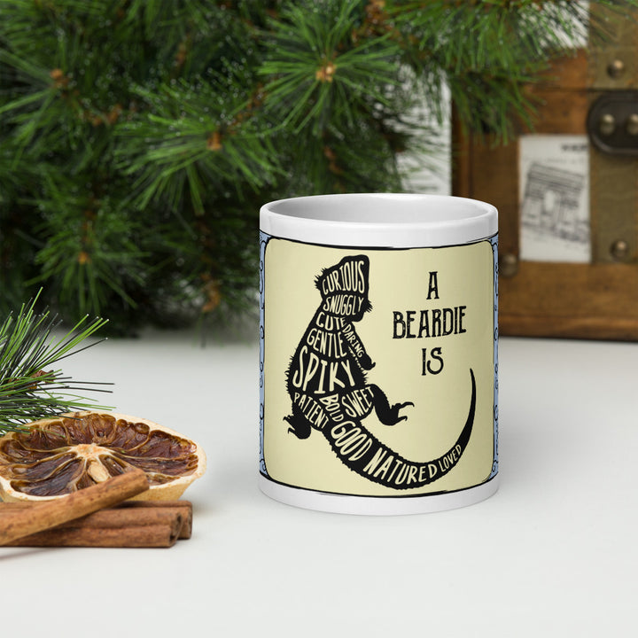 A white ceramic mug featuring a black silhouette of a bearded dragon filled with words including curious, snuggly, cute, gentle, spiky, sweet, patient, bold, and good natured. The text A BEARDIE IS is printed in black next to the lizard. The mug sits on a white table with pine branches and dried oranges on a table.