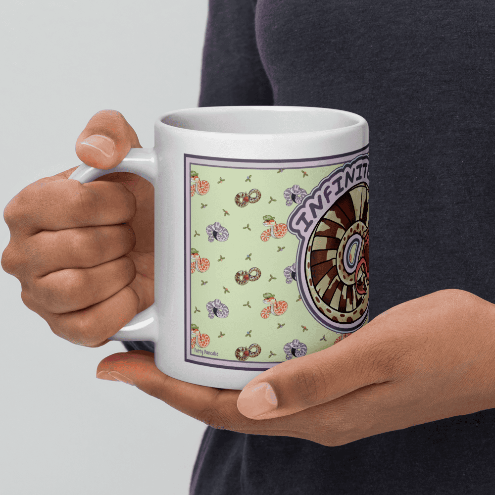 A hand cradling a white ceramic mug featuring a light green background with a repeating pattern of small, colorful snakes and plants. A circular graphic on the side displays a brown and tan wheel design with the word INFINITY printed in purple text.