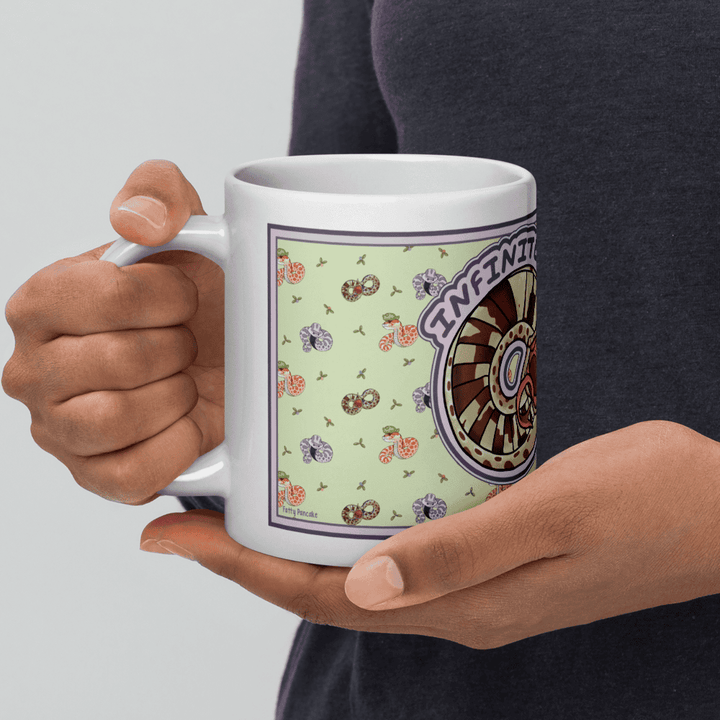 A hand cradling a white ceramic mug featuring a light green background with a repeating pattern of small, colorful snakes and plants. A circular graphic on the side displays a brown and tan wheel design with the word INFINITY printed in purple text.