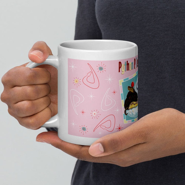 A white ceramic mug featuring a light pink background with a repeating pattern of red and white snake outlines and starburst shapes. A hand holds the handle of the mug while the other hand cups under the mugs base.