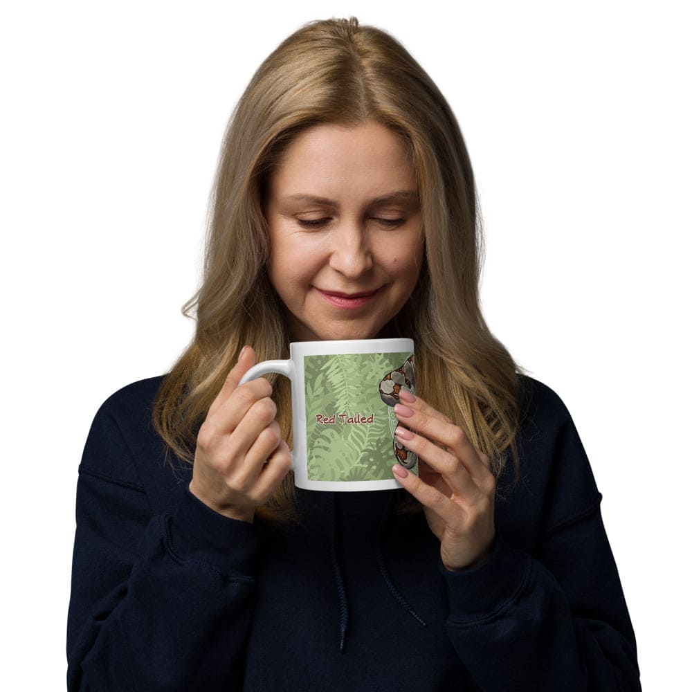 A woman holding a white ceramic mug featuring a graphic illustration of a brown and tan boa constrictor snake coiled against a green background with a fern leaf pattern. The text "Boa Constrictor" is printed in red letters on the side of the mug.