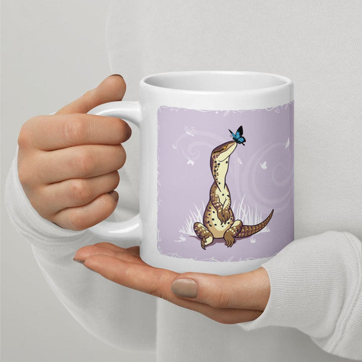 Hands holding a white ceramic mug featuring a graphic illustration of a tan spotted lizard sitting upright in grass, looking up at a small blue butterfly. The background of the illustration is a soft lavender color with white swirling accents.