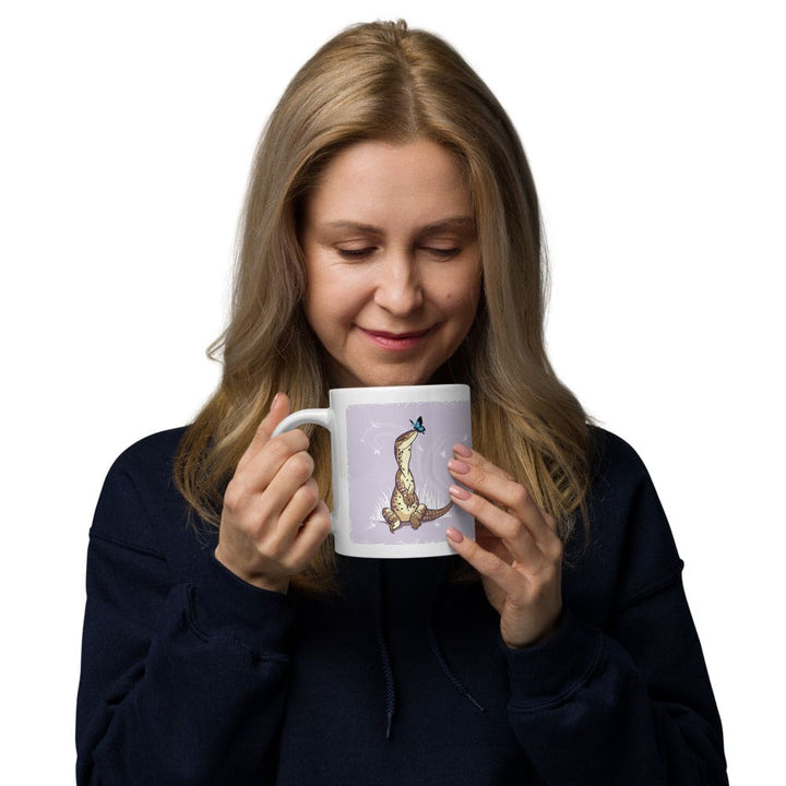A woman holding a white ceramic mug featuring a graphic illustration of a tan lizard sitting upright with a small blue butterfly perched on its nose. The background of the illustration is a soft lavender color with subtle white star-like accents.