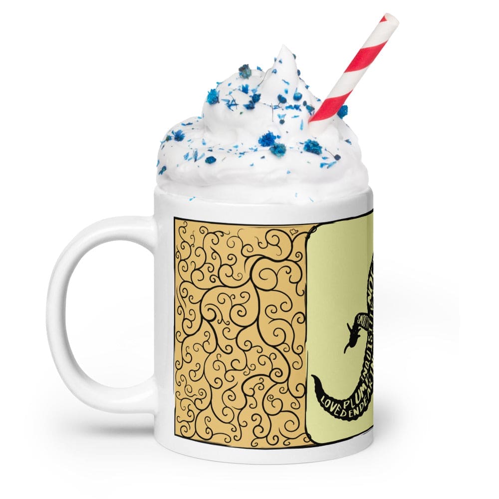 A white ceramic mug filled with whipped cream topped with blue sprinkles and a red and white striped straw. The mug features a yellow panel with black swirling filigree patterns and a black silhouette of a lizard containing text.