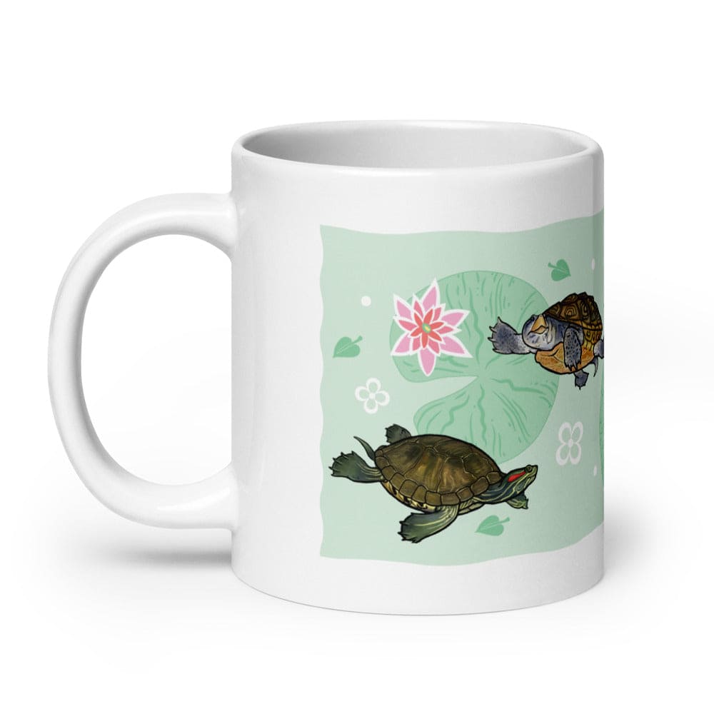 A white ceramic mug featuring a printed design of two turtles on a light green background with a pink lotus flower and lily pad motifs. The handle is on the left side of the mug.