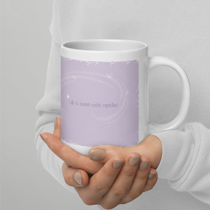 Hands holding a white ceramic mug featuring a light purple rectangular graphic on the side. The graphic contains the text "Life is sweet with reptiles" in a simple font, surrounded by faint white swirling lines and small butterfly silhouettes.