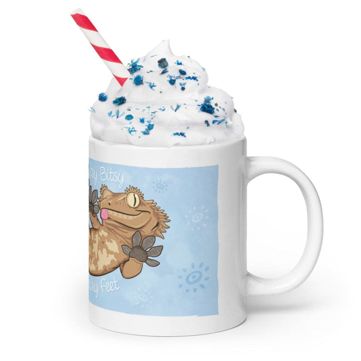  A white ceramic mug featuring a cartoon illustration of a tan crested gecko with brown spots and a pink tongue. The text reads Itsy Bitsy Sticky Feet. The mug is topped with white whipped cream, blue sprinkles, and a red and white striped straw.