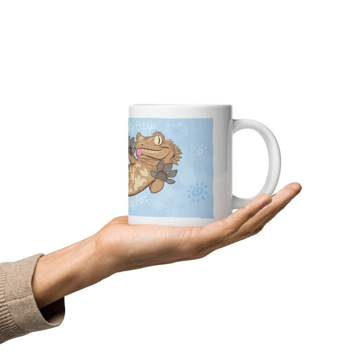 A hand holding a mug in the palm that shows a cartoon crested gecko.
