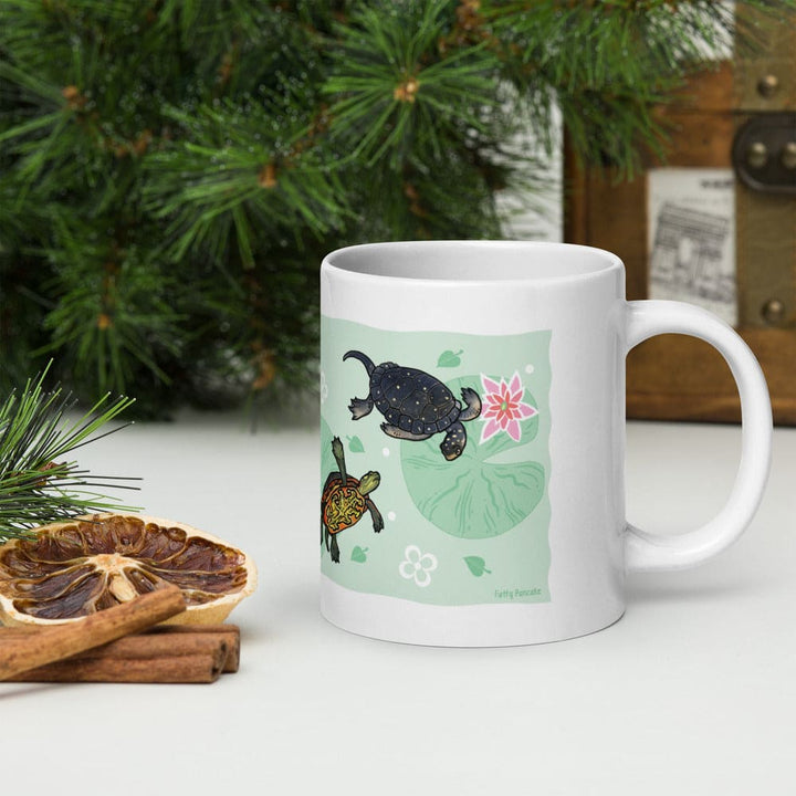 A white ceramic mug with a light green illustration of two turtles, a pink flower, and lily pad leaves, set on a white tabletop with pine branches, cinnamon sticks, and a dried orange slice in the foreground.