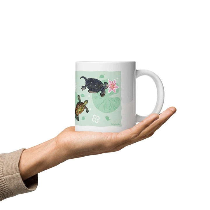 A hand holding a white ceramic mug with a light green illustration of fresh water turtles, lily pads, and a pink starfish on a plain white background.