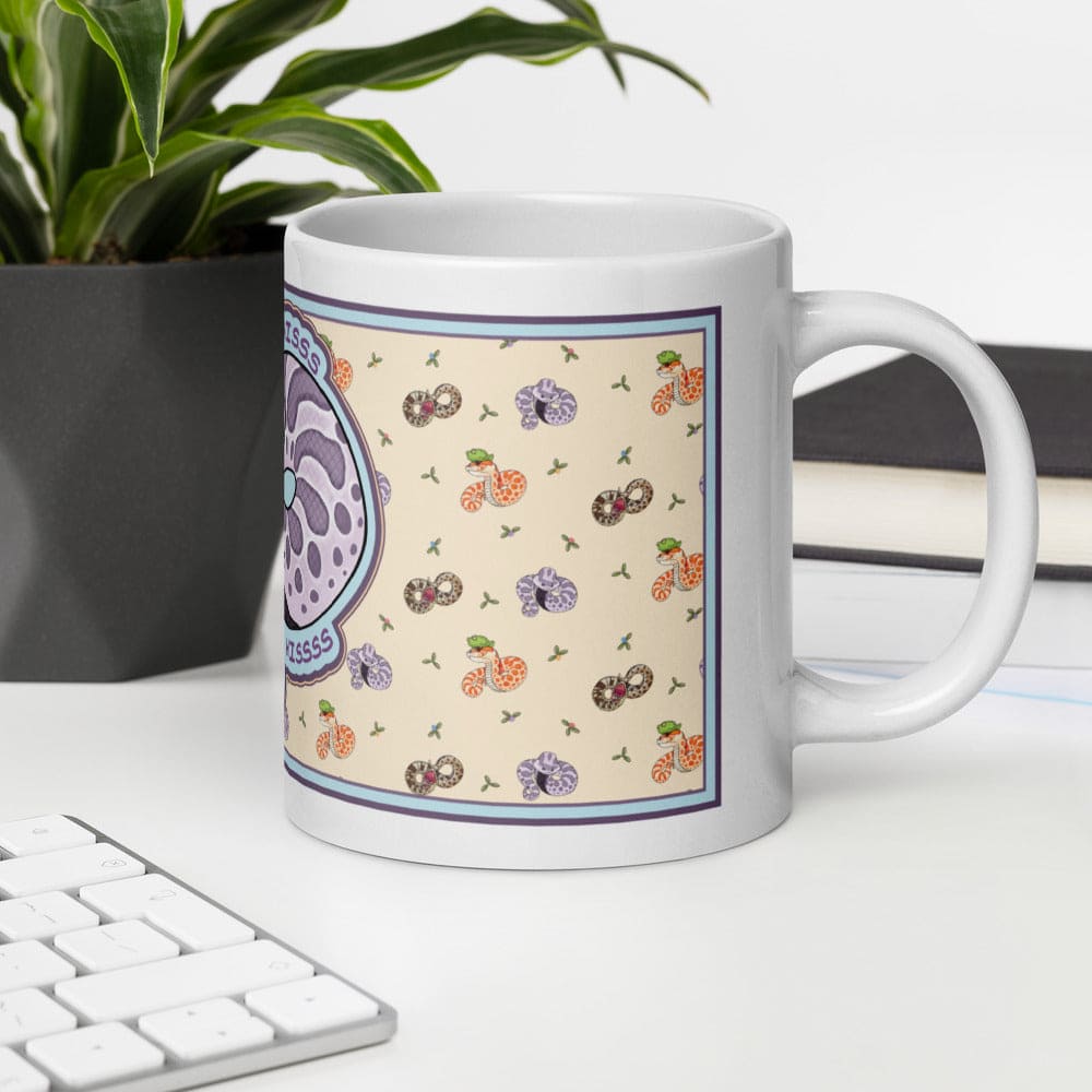 A white ceramic mug featuring a repeating pattern of small snakes in orange, purple, and brown on a cream background. A larger circular graphic on the left side displays a purple snake with the text "HISSS" and "HISSSSSS" in blue. The mug sits on a white table with a keyboard and potted plant.