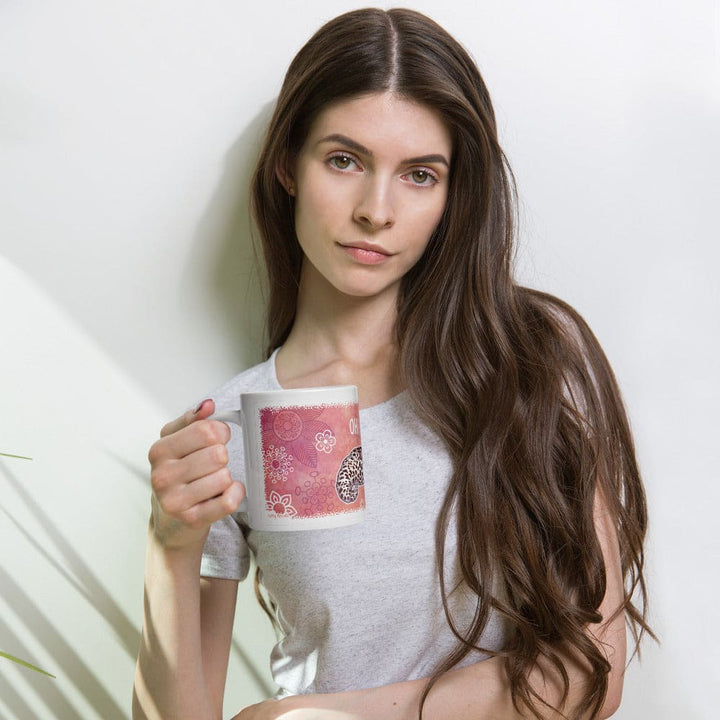 A woman with long brown hair holds a white ceramic mug with a pink and white floral pattern. She is wearing a light gray t-shirt and posing against a white wall with soft shadows.