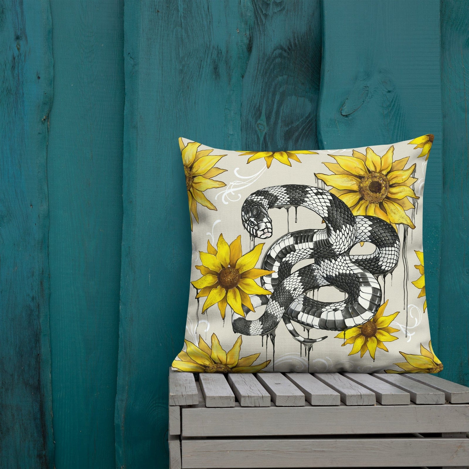 A square-shaped throw pillow with a pattern of a kingsnake intertwined with sunflowers, predominantly in yellow and black colors, placed on a wooden surface with a blue wall in the background. Pillow measures 22 inch x 22 inch.